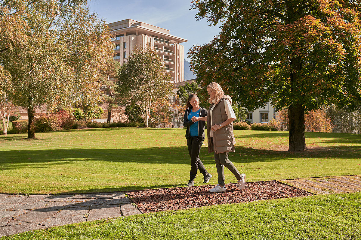 Zwei Frauen spazieren und unterhalten sich in einem sonnigen Park, umgeben von grünem Gras und hohen Bäumen. Ein modernes Gebäude im Hintergrund verleiht dem Bild einen urbanen Touch.