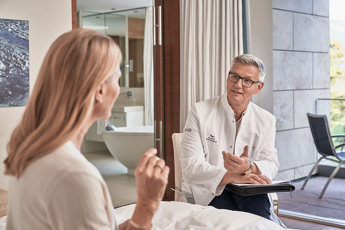 Ein Arzt im weißen Kittel berät sich mit einem Patienten in einem hellen Raum, der eine ruhige und professionelle Atmosphäre schafft. Im Hintergrund ist ein modernes Badezimmer zu sehen.